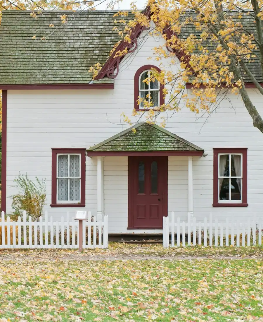 White clapboard house with a red door and a picket fence under autumn maples